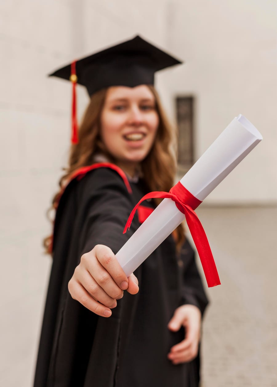 A graduate smiling and holding a rolled diploma tied with a red ribbon
